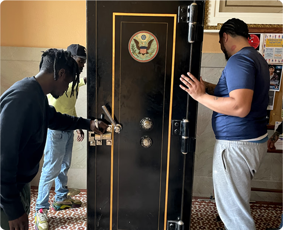 Two people opening a large black safe door in a room with a patterned floor and wall emblem.