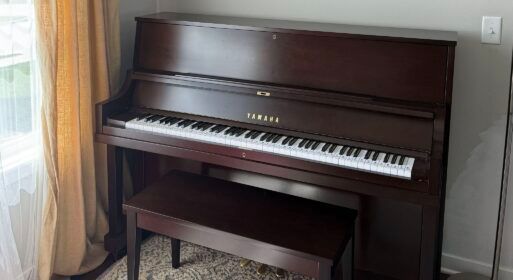 Dark wood upright piano with matching bench in a living room by a window with curtains.