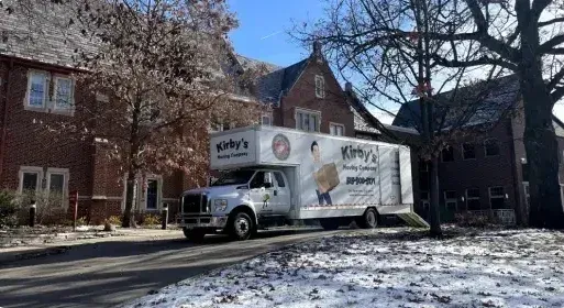 White box truck parked on a snowy street beside brick buildings and leafless trees.