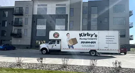 Moving truck parked in front of a gray apartment building on a sunny street.