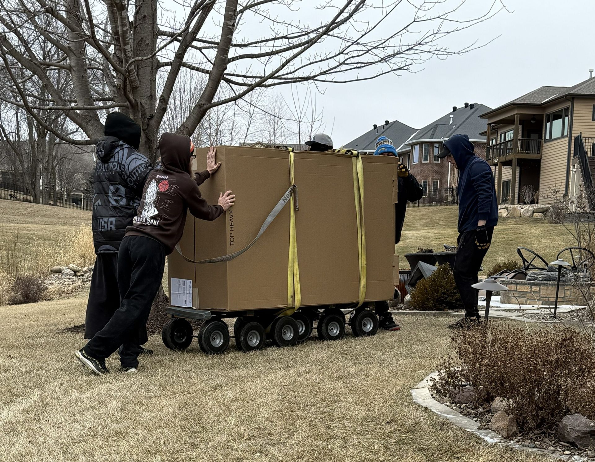 Three people push a cardboard structure on wheels across a suburban lawn