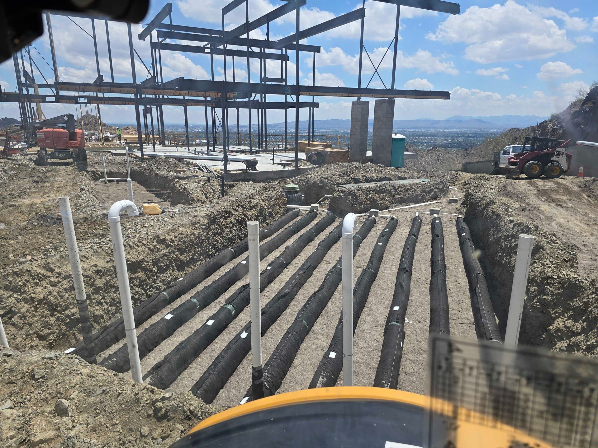 Construction site with excavated trenches, pipes, and a building frame. Heavy machinery visible in the background.