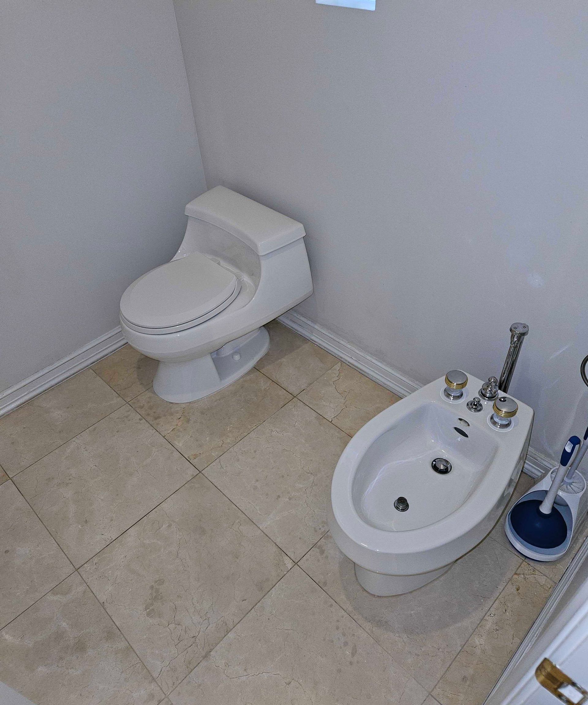 Small bathroom with toilet, bidet, beige tile floor, and white walls.