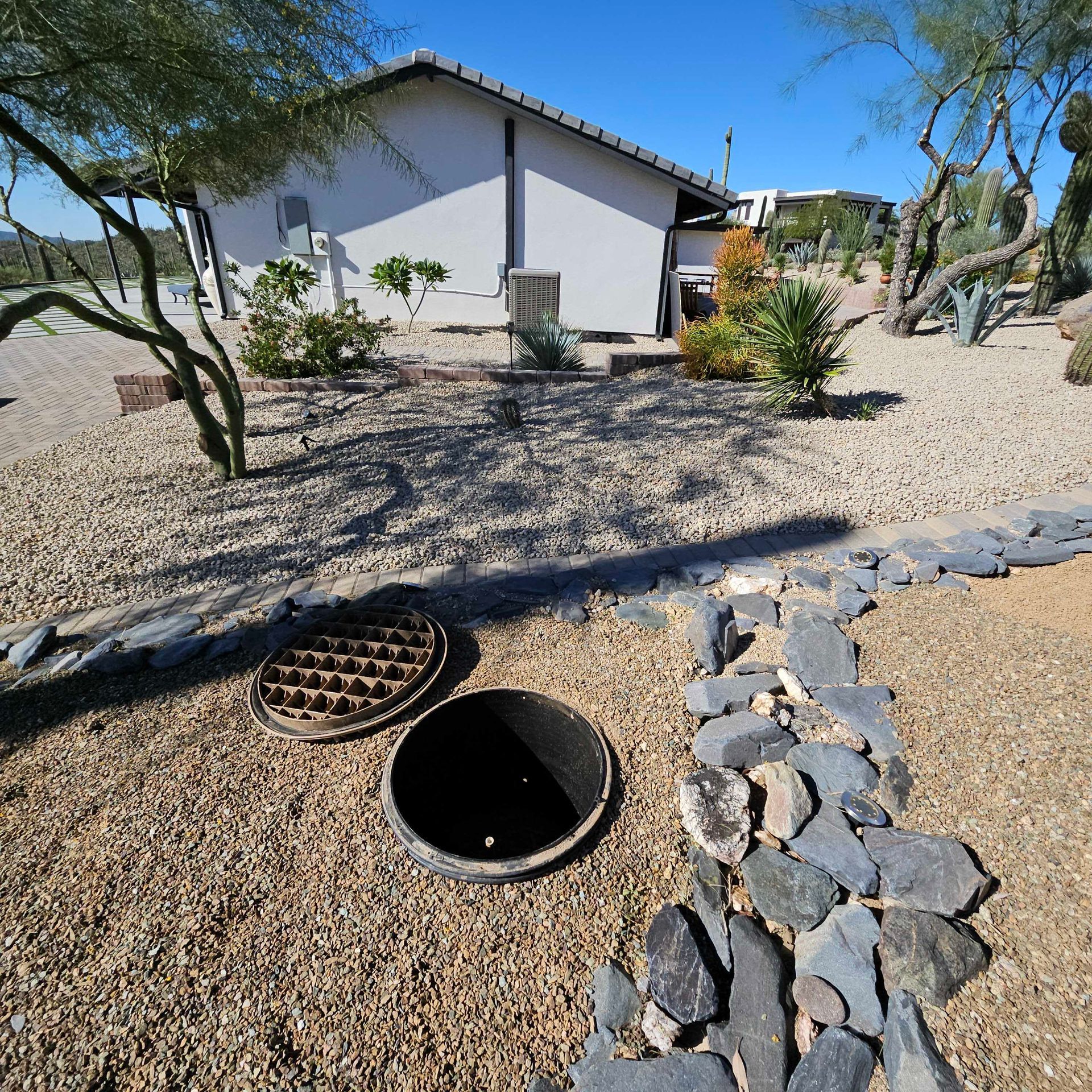 Desert yard with a white house, gravel landscaping, trees, and two open utility access covers in the foreground