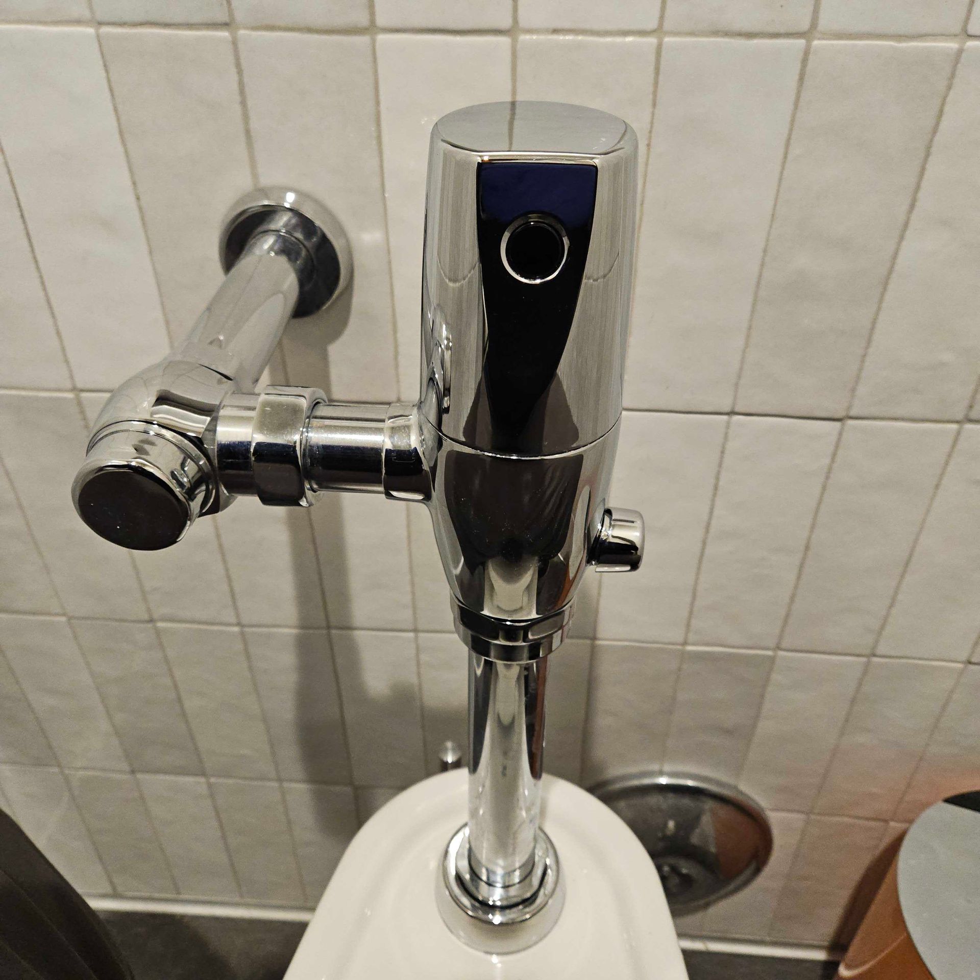 Chrome flushometer on a white wall-mounted urinal in a tiled restroom
