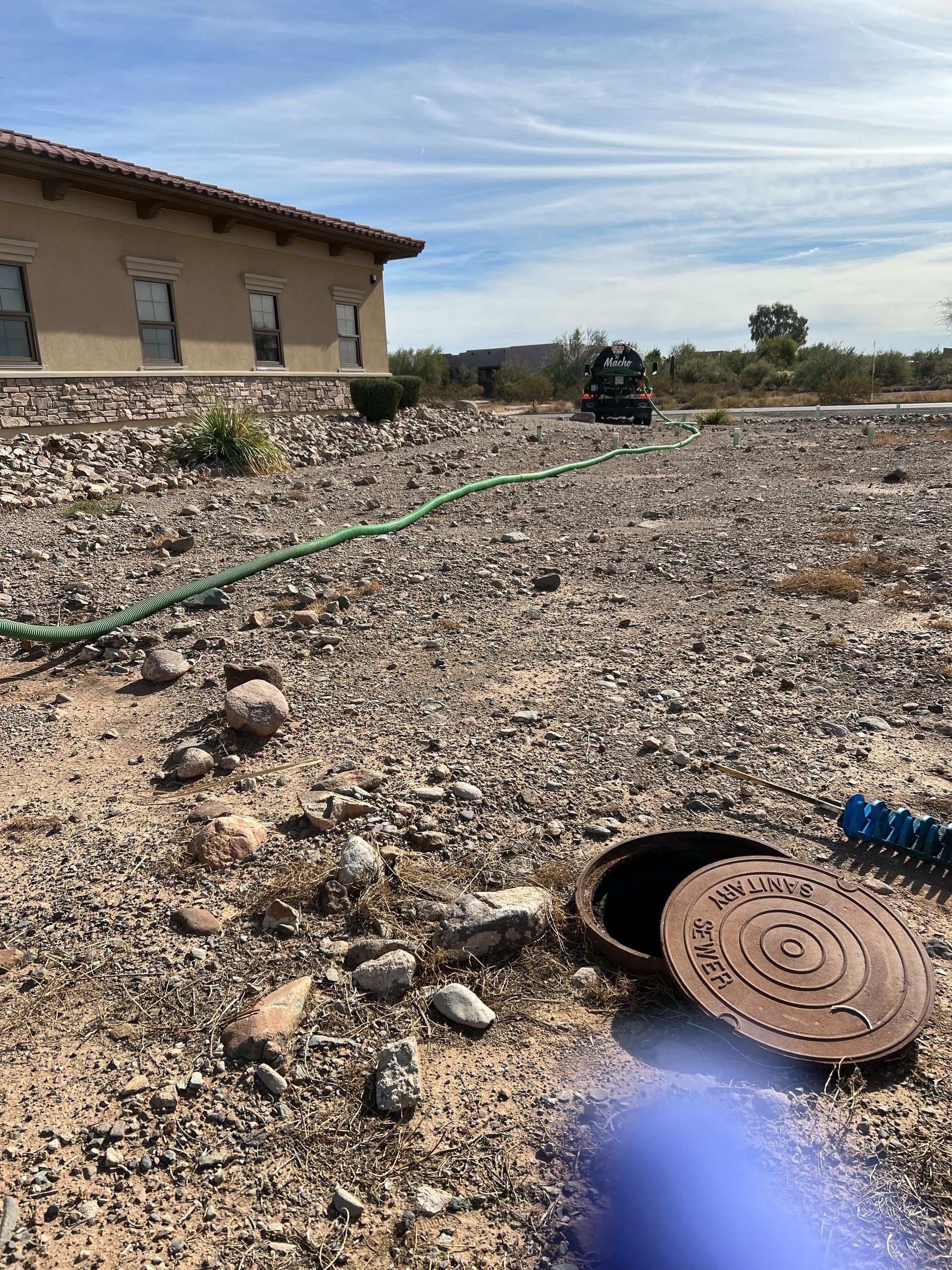 Rubble-strewn lot beside a house, with a green hose and a rusted manhole cover in the foreground.