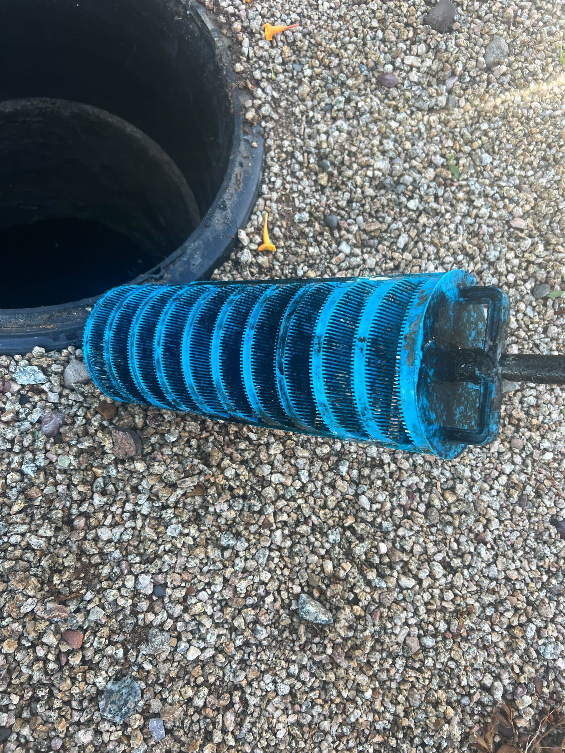 Blue-and-black striped filter cartridge beside a drain opening on gravel.