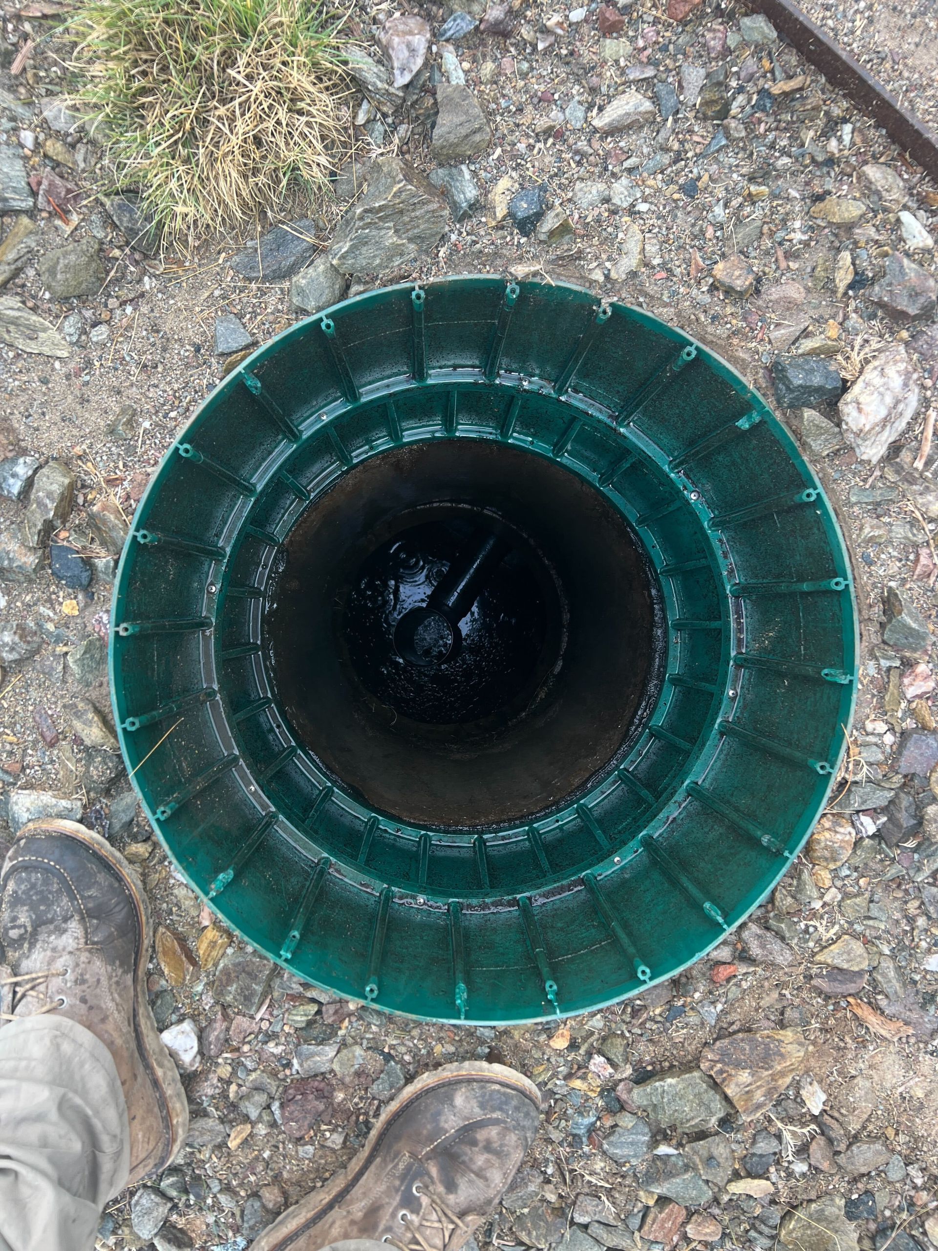 Top view of a green-rimmed water well opening in rocky ground, with boots visible at the edge.