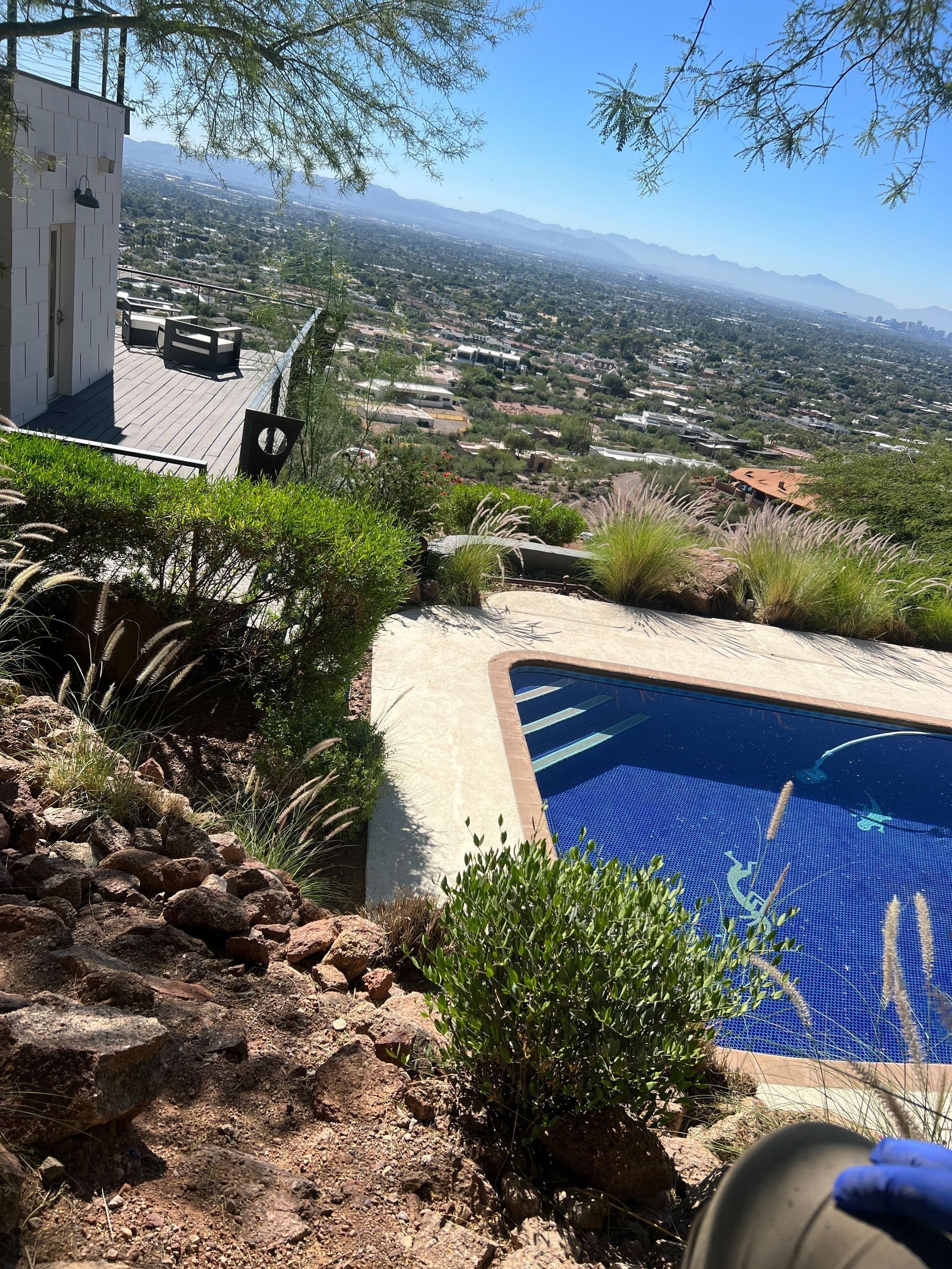 Blue-tiled pool overlooking a sunlit hillside cityscape with shrubs and a patio area