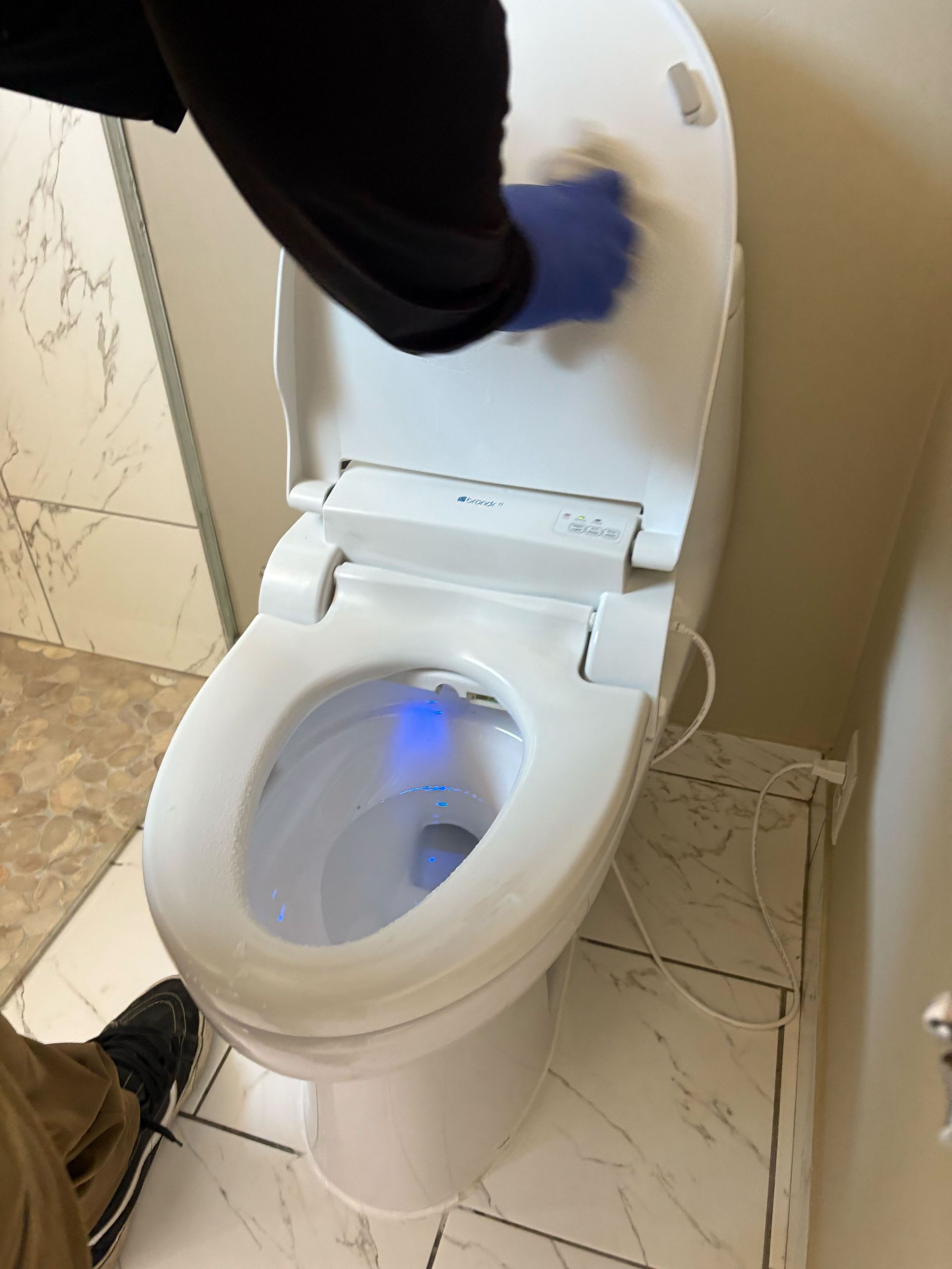 Open toilet with a hand cleaning the lid, blue light inside the bowl, in a small bathroom.