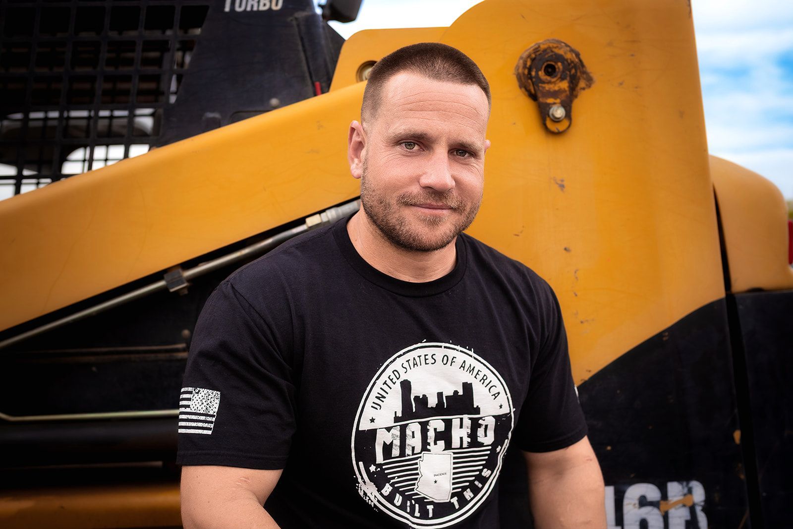 A man with in a black shirt is standing in front of an excavator.