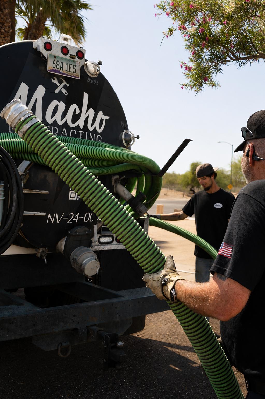 Two people handle green hoses at the rear of a black truck labelled Macho, outdoors in daylight.