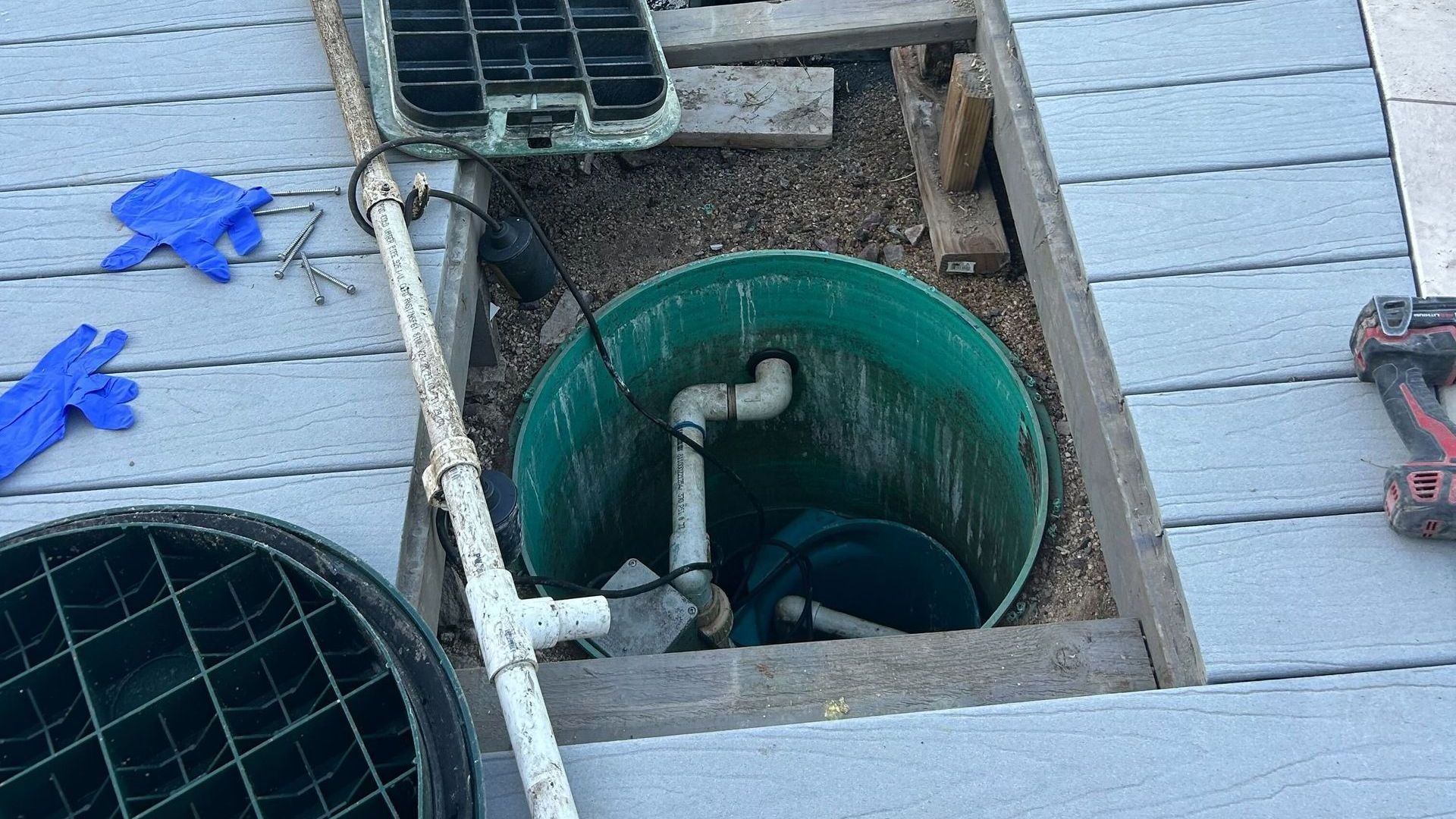 Open drainage pit with a green utility barrel and pipes under a deck, with tools nearby
