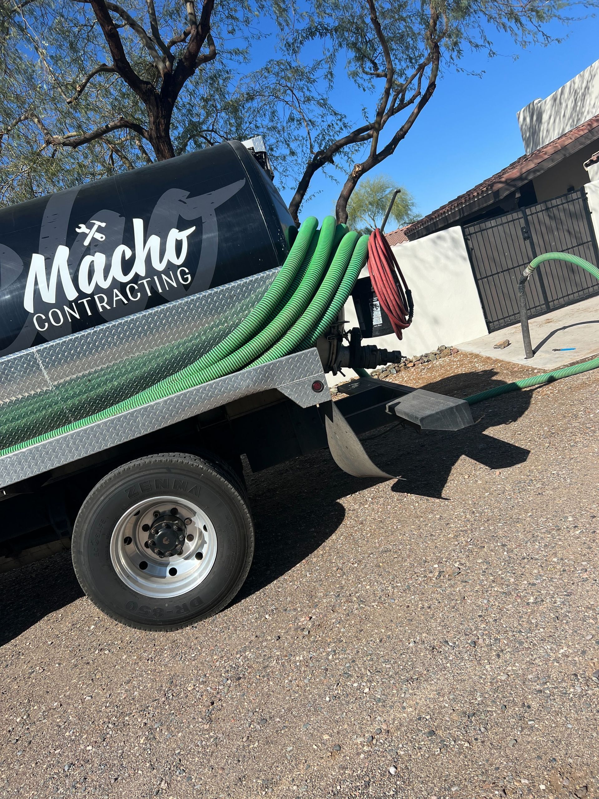 Black utility trailer with green hoses and Macho Painting logo parked on a gravel driveway under a blue sky