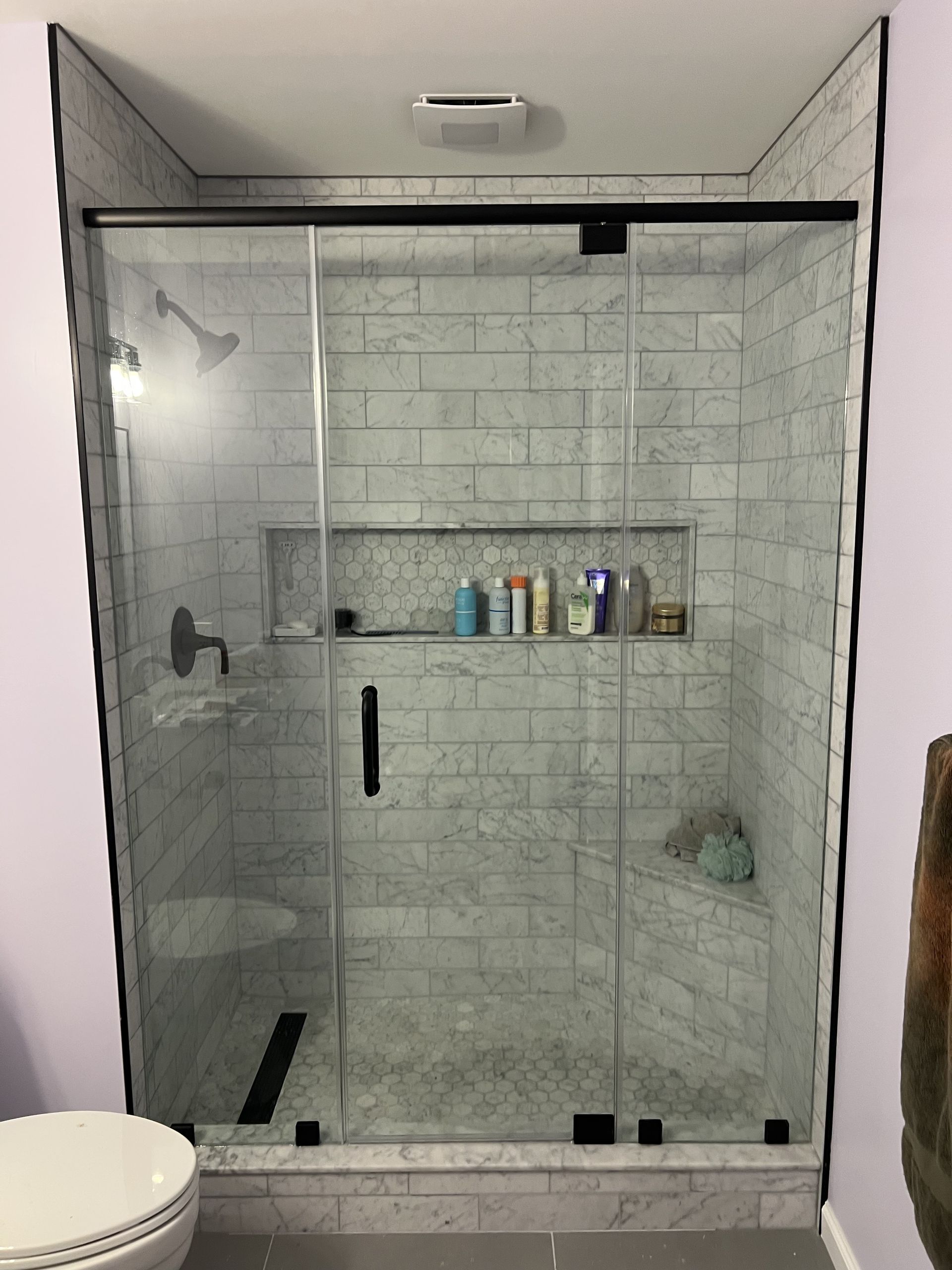 A modern glass-enclosed shower with white marble-style subway tiles, a built-in shelf, and black fixtures.