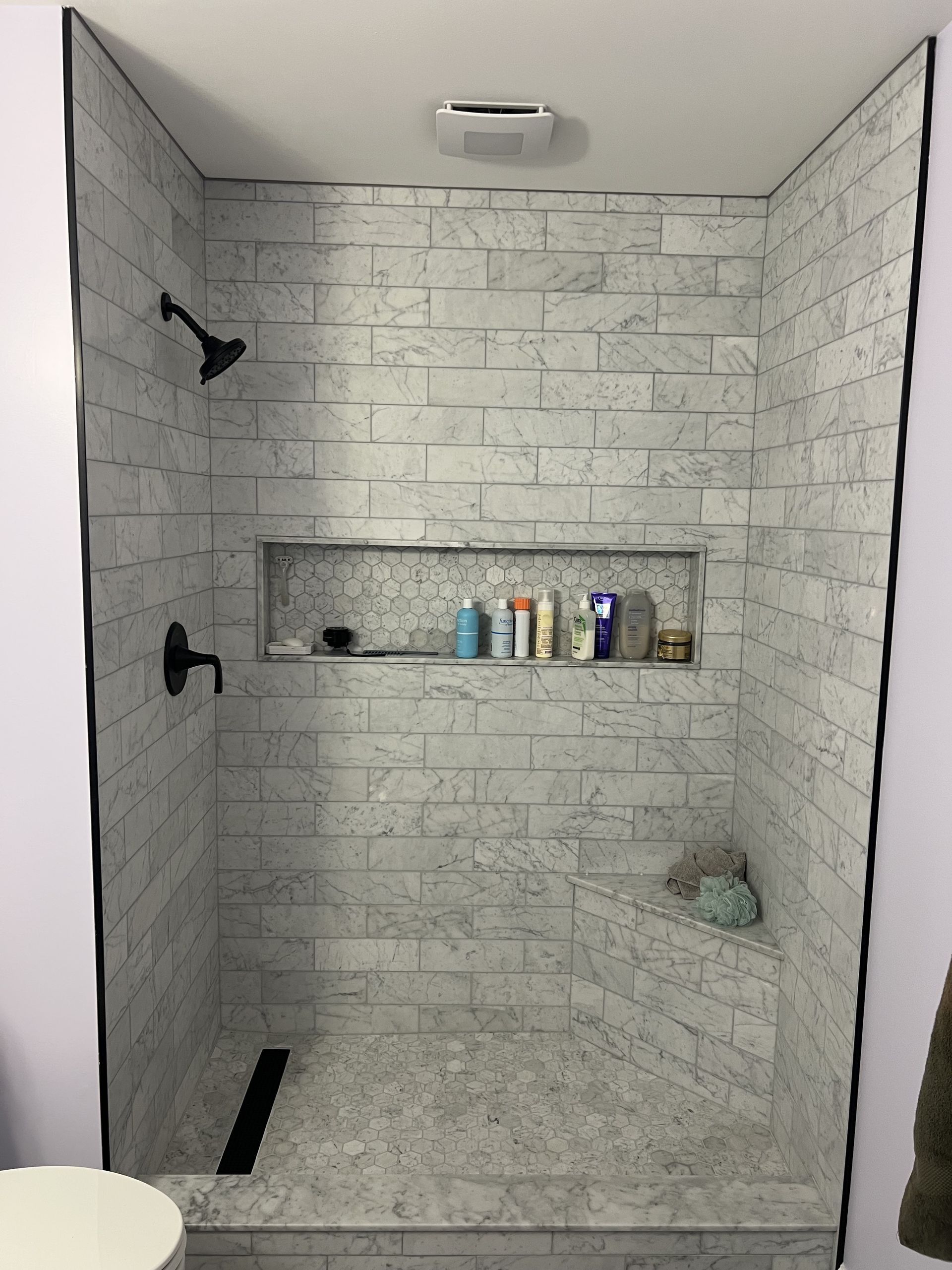 A shower stall with light gray brick-pattern tiles, a built-in recessed shelf with toiletries, a corner seat, and a drain.