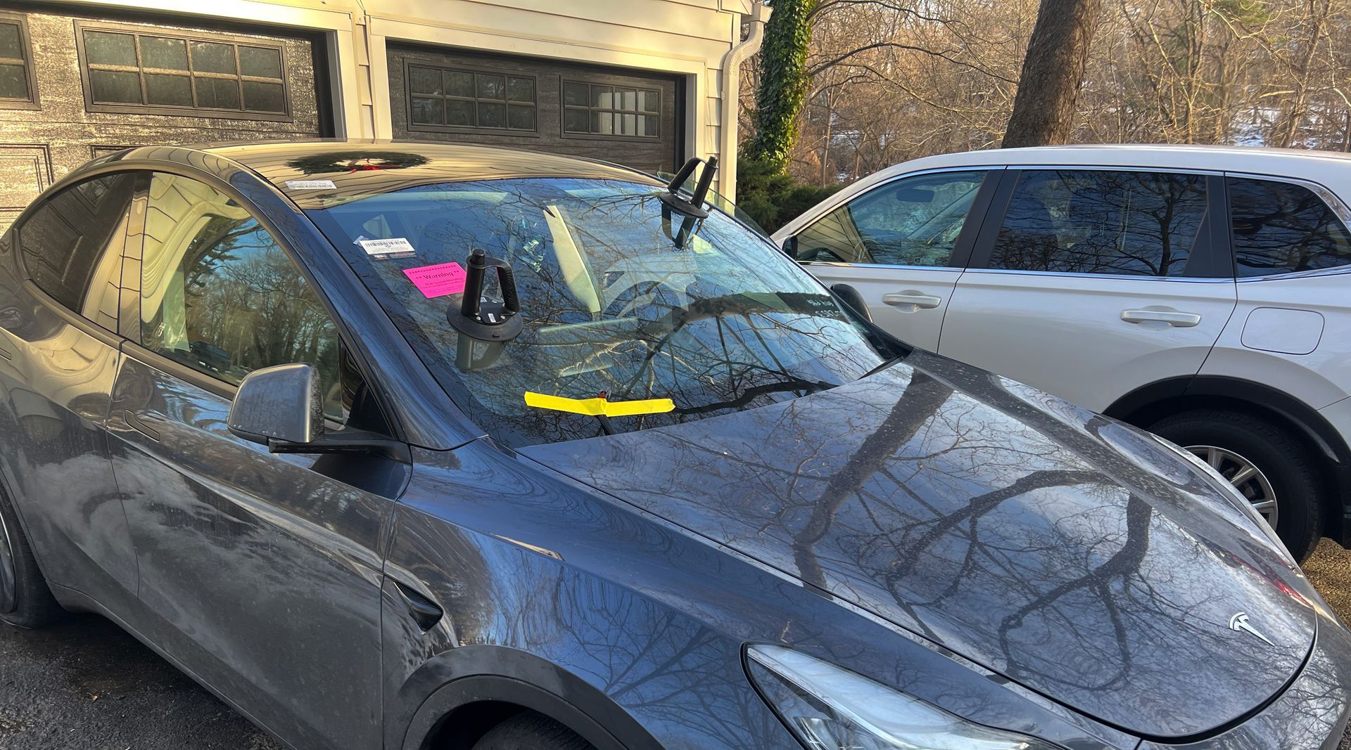 Dark gray Tesla Model Y parked, windshield wipers up, next to white SUV in front of a garage.