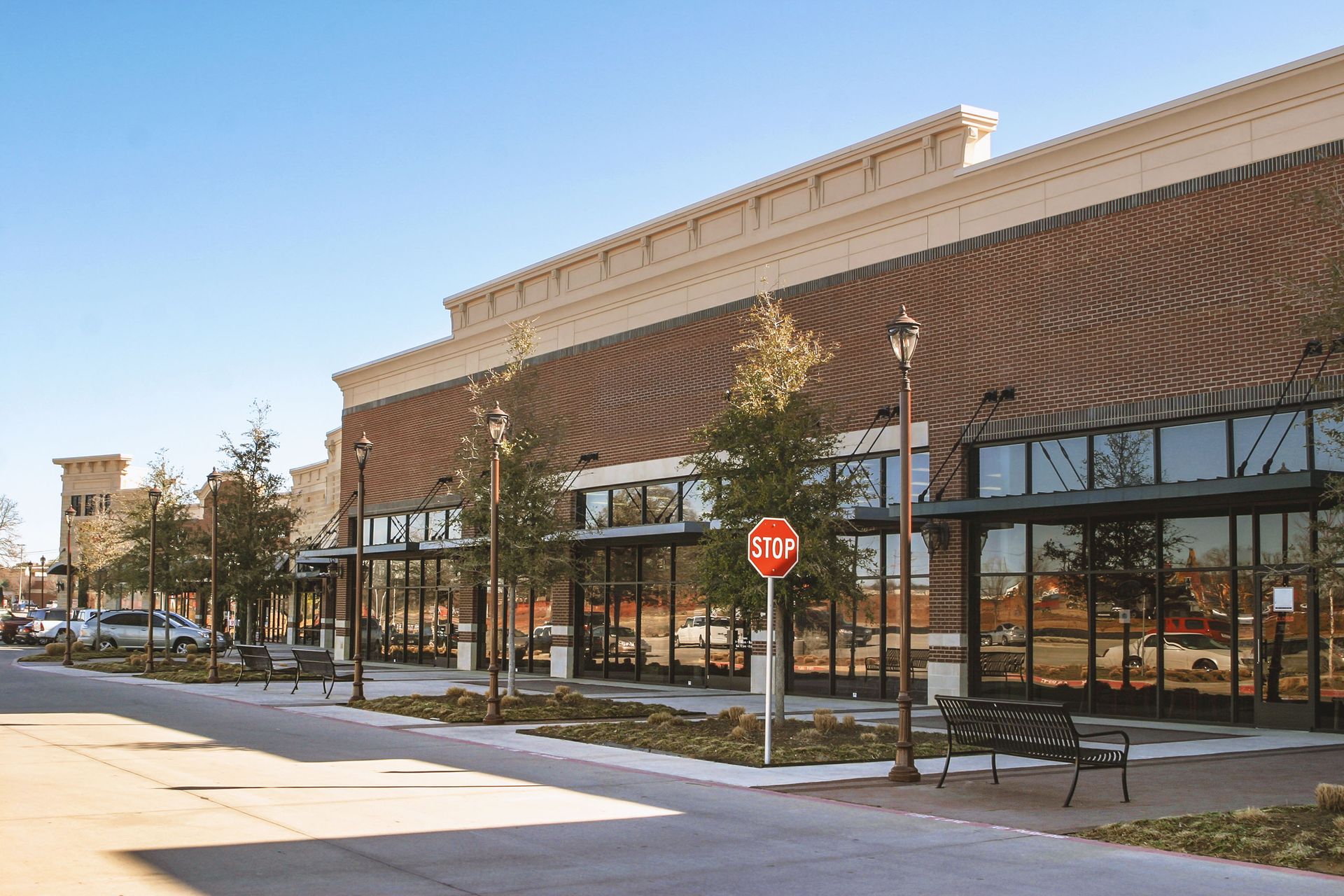 Row of brick storefronts with glass windows, sidewalk, trees, and benches. A stop sign is visible.