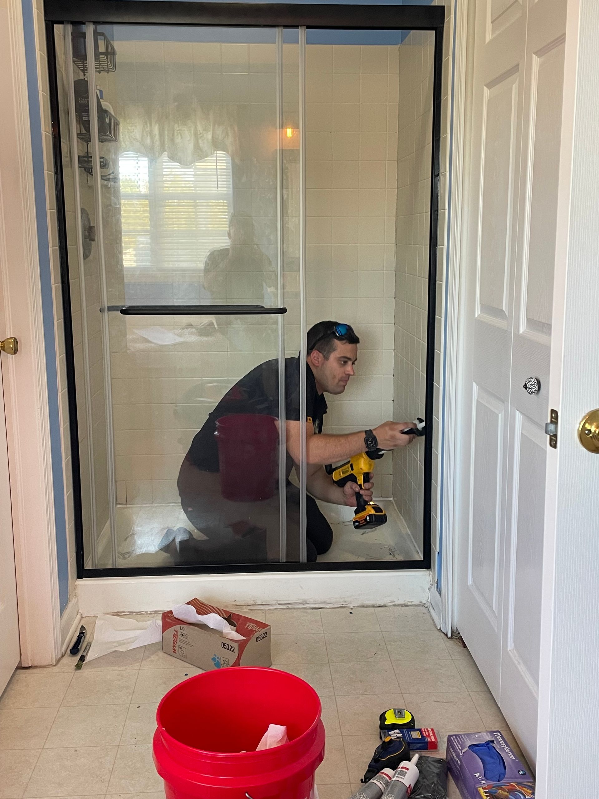 A person installing a shower door. They are kneeling, using a drill inside the shower.