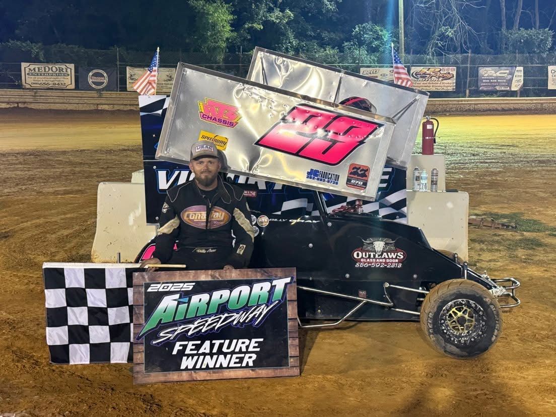 Race car driver in black suit stands with winning race car. Airport Speedway Feature Winner sign in front.