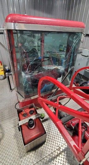 Damaged red and silver tractor cab with shattered windshield, metal frame, and fuel tank.