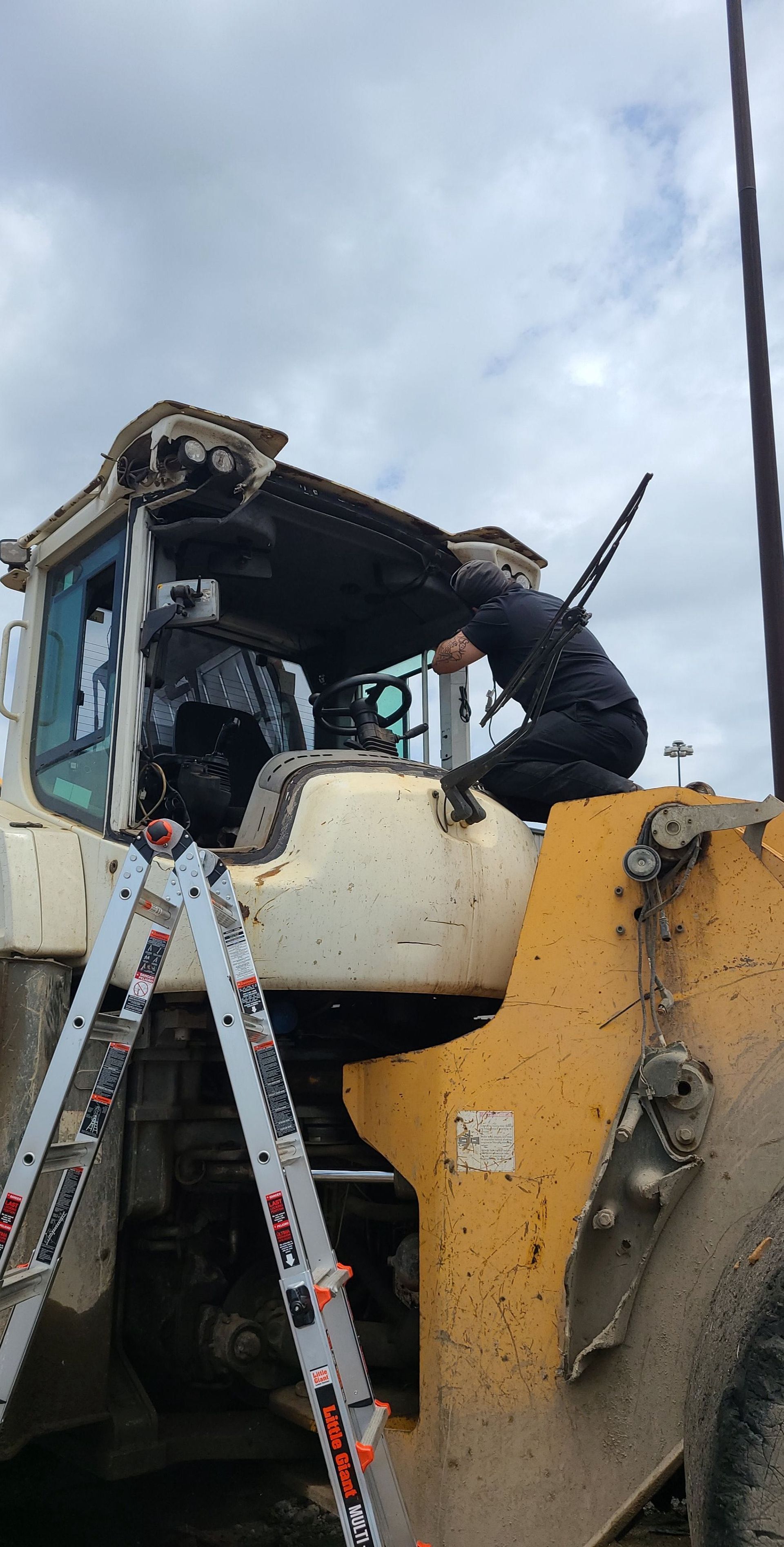 A person works on the roof of a large construction vehicle, using a ladder. Overcast sky.