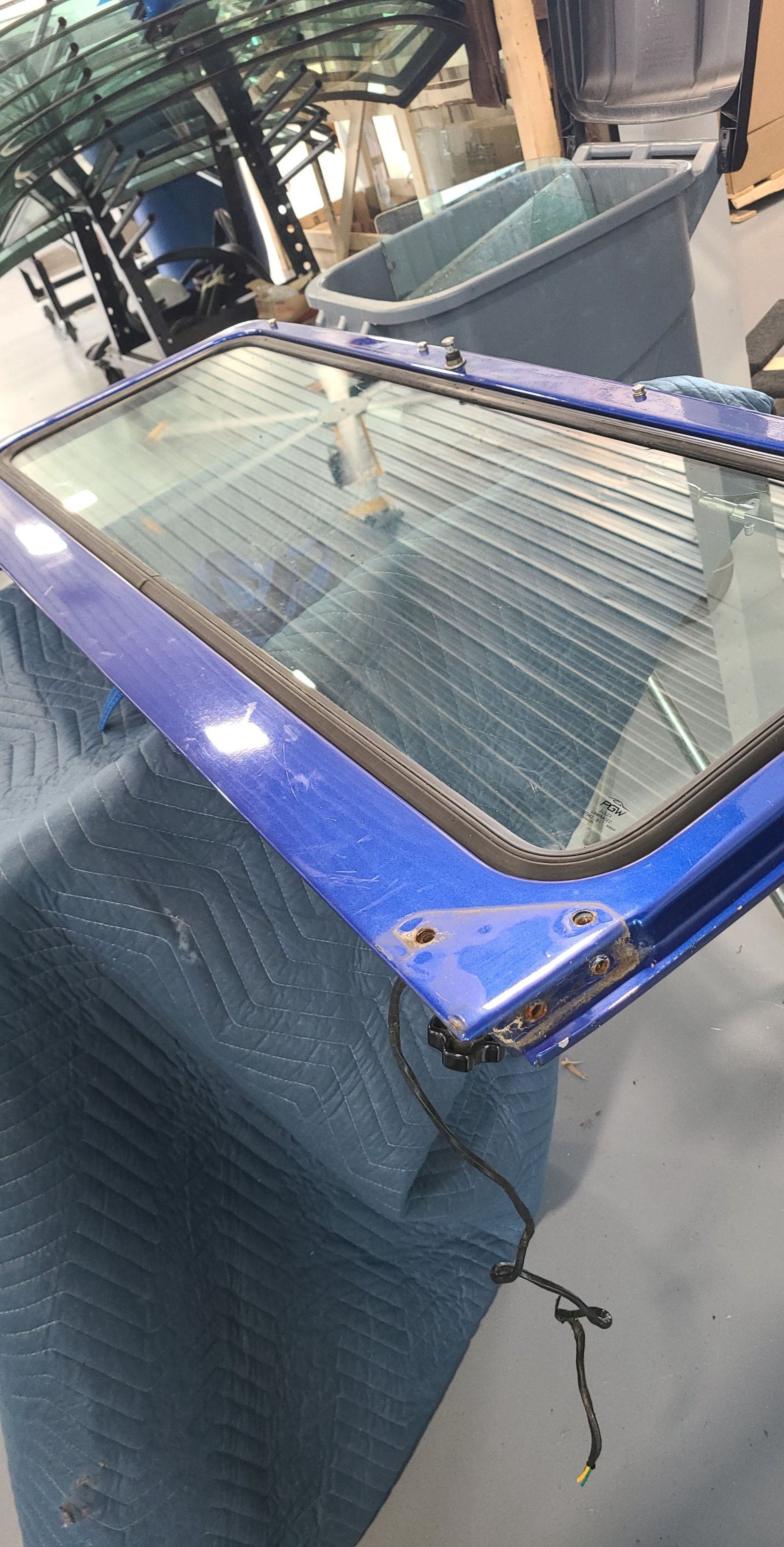Blue car window frame with glass, lying on a blue padded surface; a large plastic container is in the background.