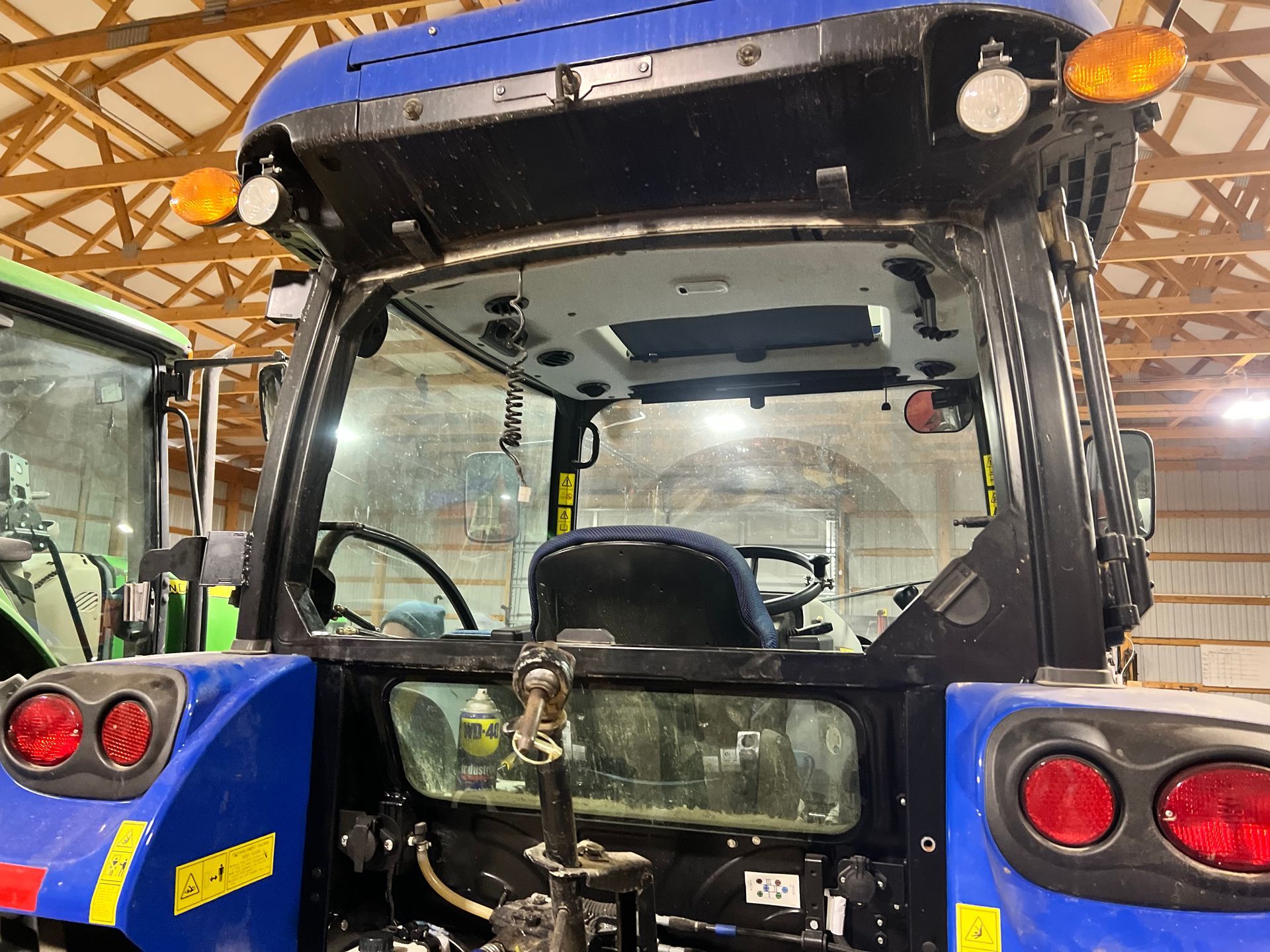 Blue tractor cab with glass windows, inside controls visible, rear lights, and a green tractor partially visible.