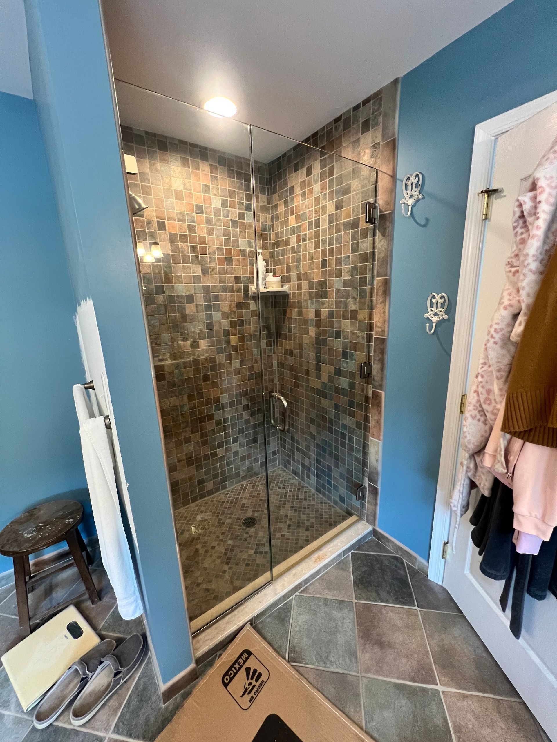 A tiled shower with glass doors and blue walls. Coats hang on the door. Wooden stool and shoes on the floor.