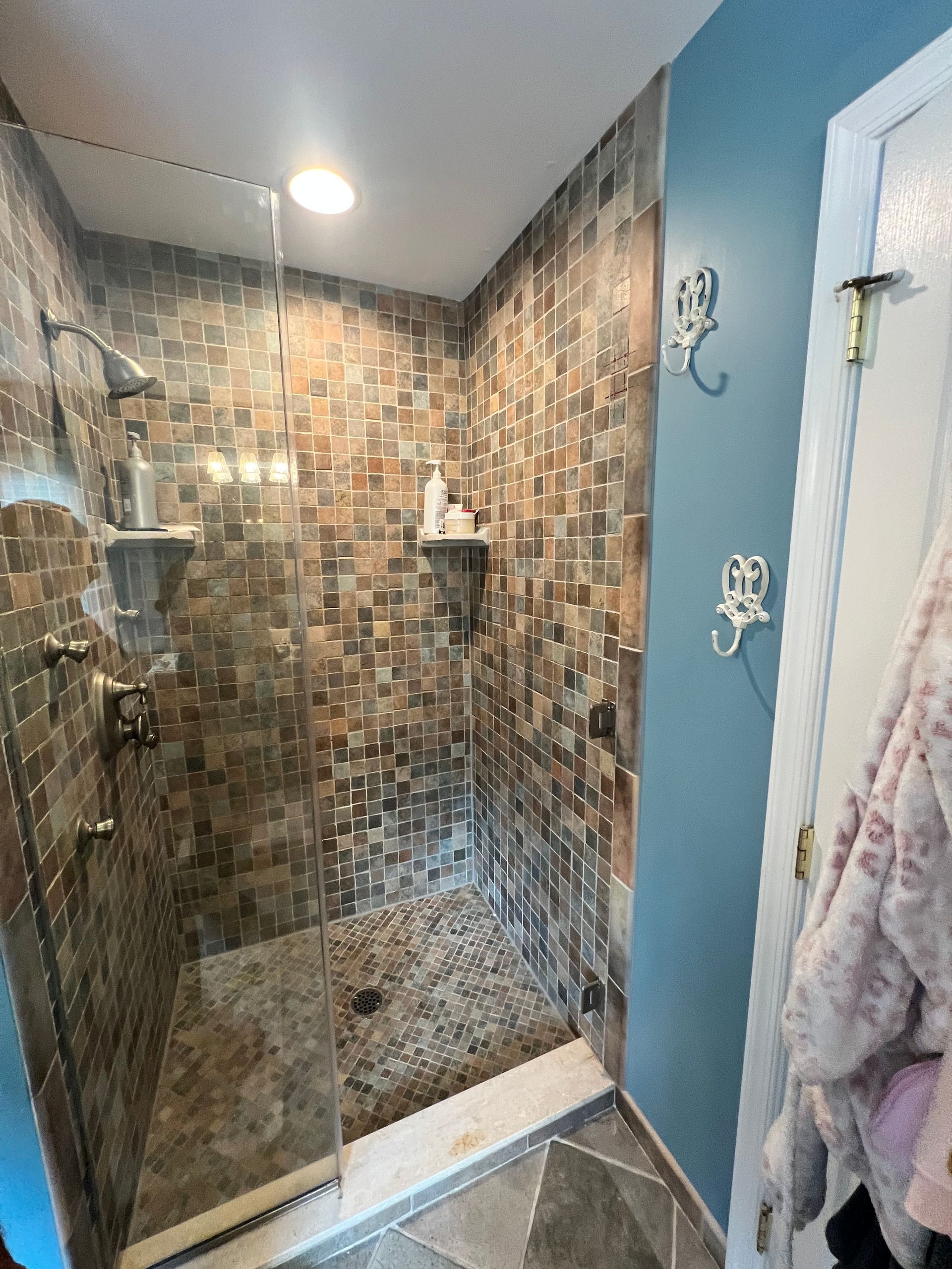 Shower with stone tile walls, glass door, pebble floor, and blue wall with hooks.