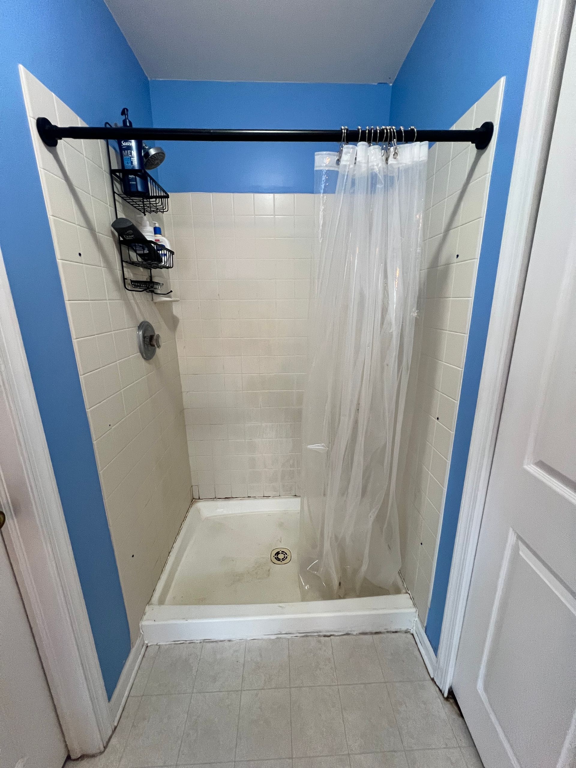 Small shower stall with blue walls, cream tile, and a clear shower curtain.