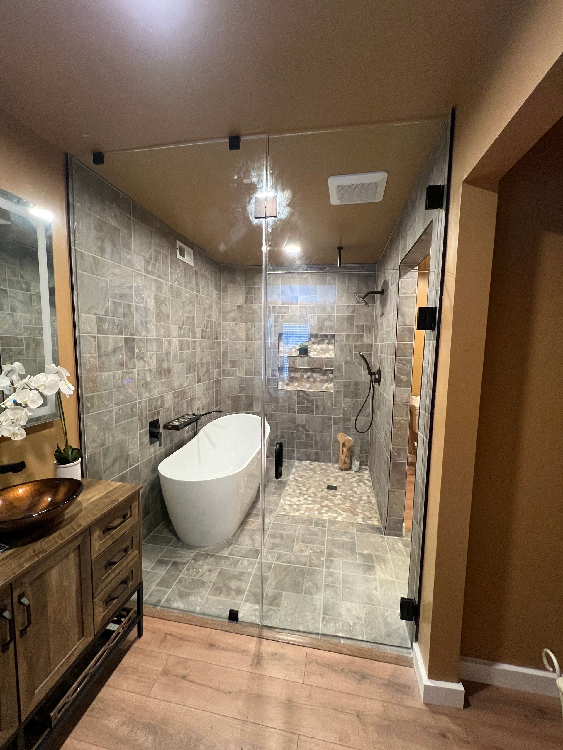 Modern bathroom with a glass-enclosed shower, freestanding tub, stone tile, and wooden vanity.