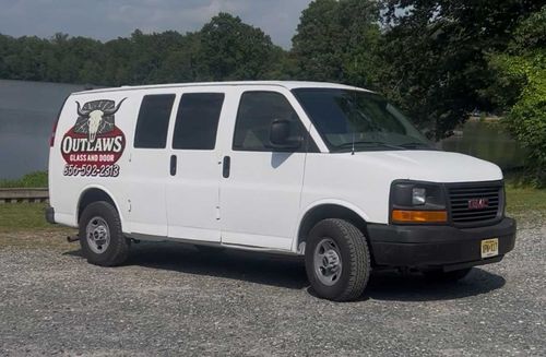 White GMC van with "Outlaws BBQ & Meats" logo parked outdoors near water and trees.