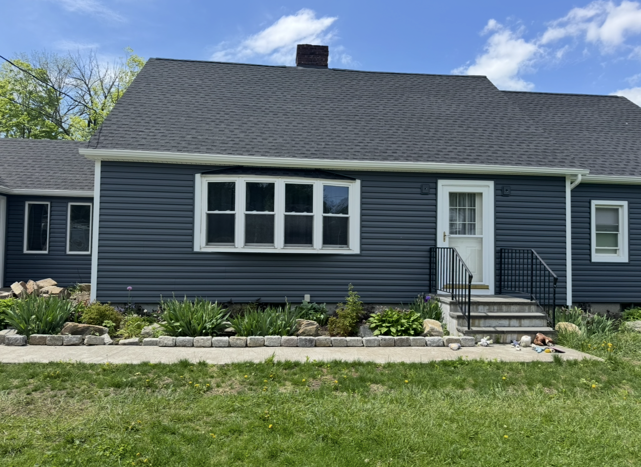 A blue house with a gray roof and white windows is sitting on top of a lush green field.