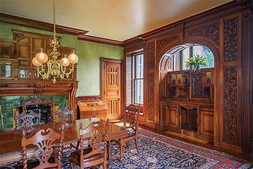 Ornate wooden dining room with fireplace, green walls, chandelier, dark wood furniture, and patterned rug.