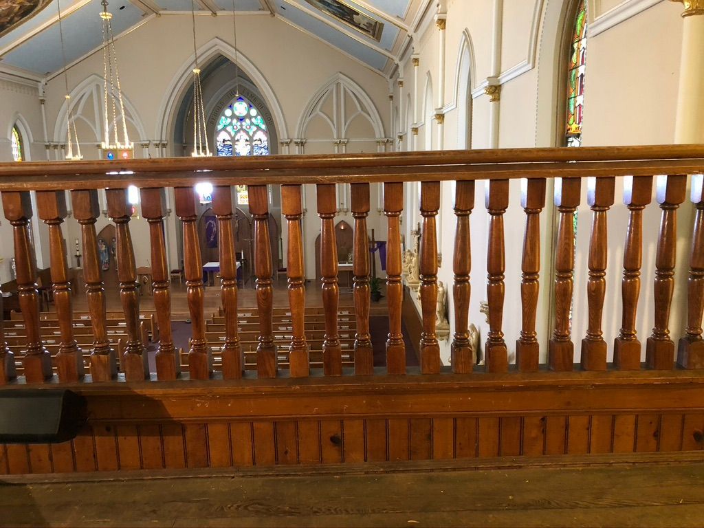 Wooden railing inside a church, overlooking pews and a stained-glass window.