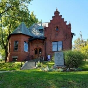 Red brick historic building with a turret and gabled roof, on a grassy lawn.