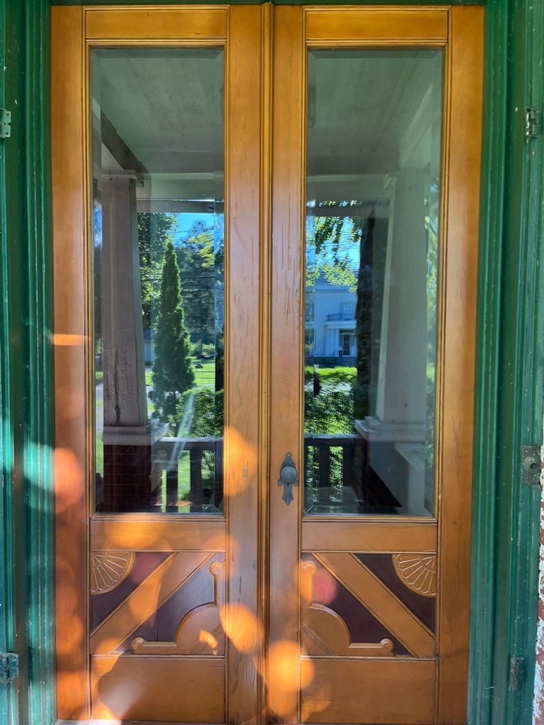 Double wooden doors with glass panes, reflecting a porch and foliage. Green trim, decorative bottom panels.