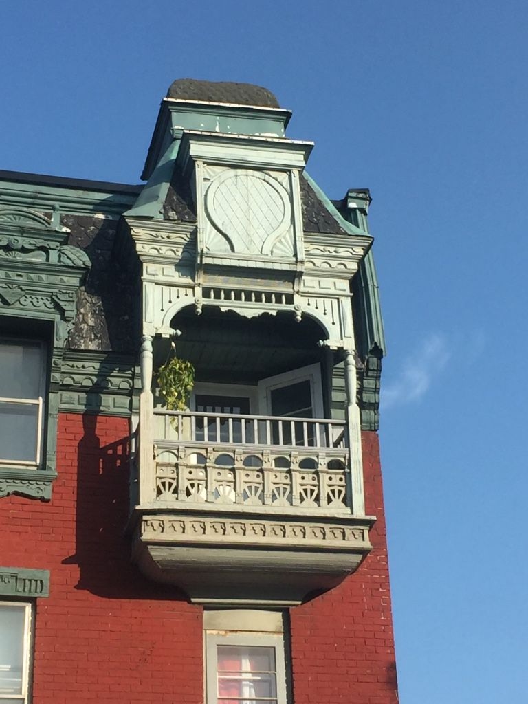 Ornate, light blue balcony with detailed trim on a red brick building, against a blue sky.
