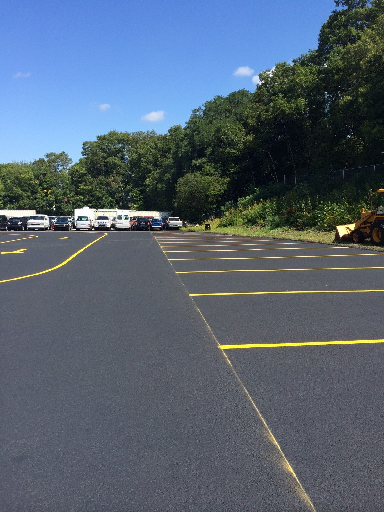 Empty asphalt lot with yellow lines, parked vehicles in the distance, and trees under a blue sky