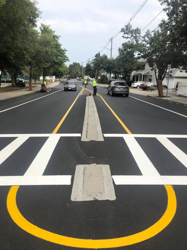 Street with raised pedestrian median, painted crosswalk, and worker in yellow vest managing traffic.