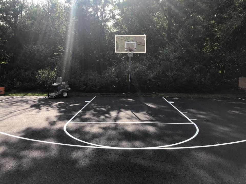 Outdoor basketball court with hoop, white boundary lines, and trees in the background.