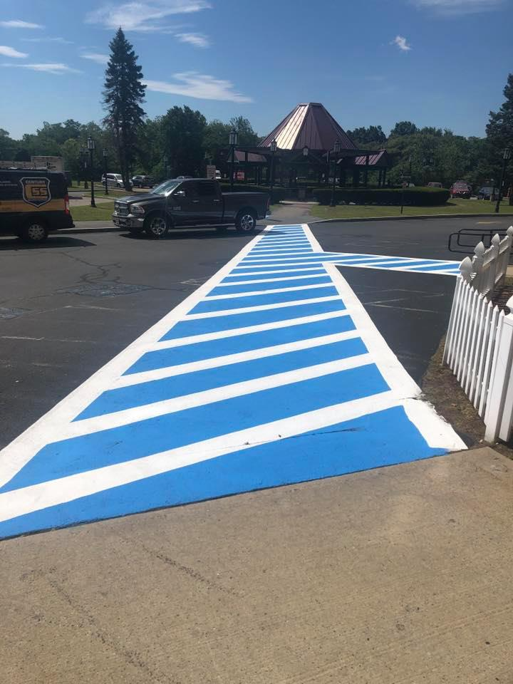 Blue-and-white striped pedestrian ramp leading across a parking lot toward a building and trees