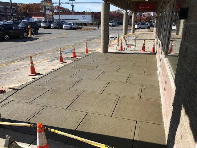 Sidewalk under construction beside a building, with orange cones and caution tape blocking the walkway.