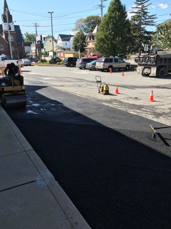 Street work area with orange cones, equipment, parked vehicles, and houses along a sunny road.
