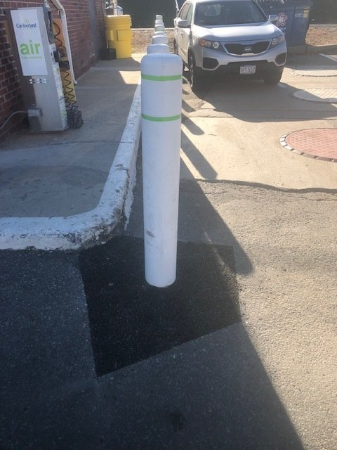 White bollard on a sidewalk beside a road, with a parked SUV and storefronts in the background.