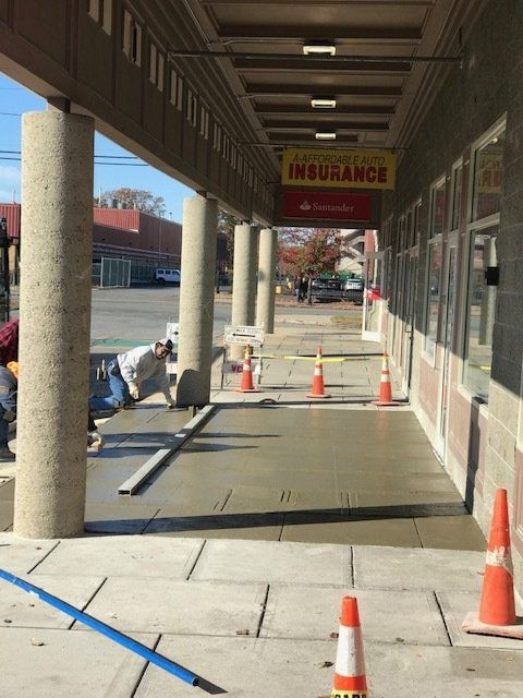 Covered storefront sidewalk with orange cones and a yellow “NO BARRIERS” sign by a closed construction area