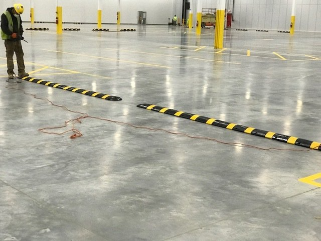 Empty industrial warehouse floor with black-yellow barriers and a worker standing at left.