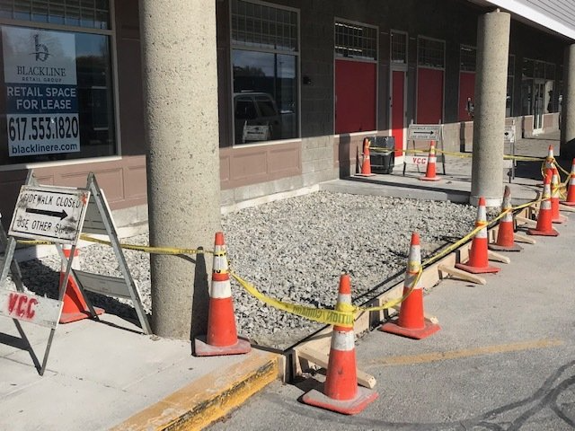Sidewalk construction area blocked by orange cones and caution tape outside a storefront.
