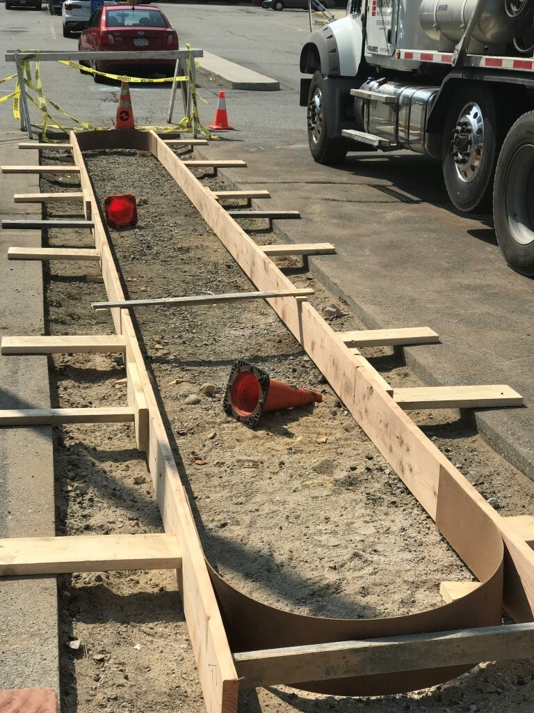 Construction workers laying forms and rebar for a curved concrete curb in a street lane, with trucks nearby